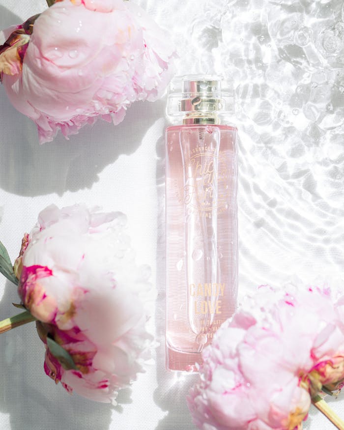 A delicate perfume bottle surrounded by pink peonies under sunlight.