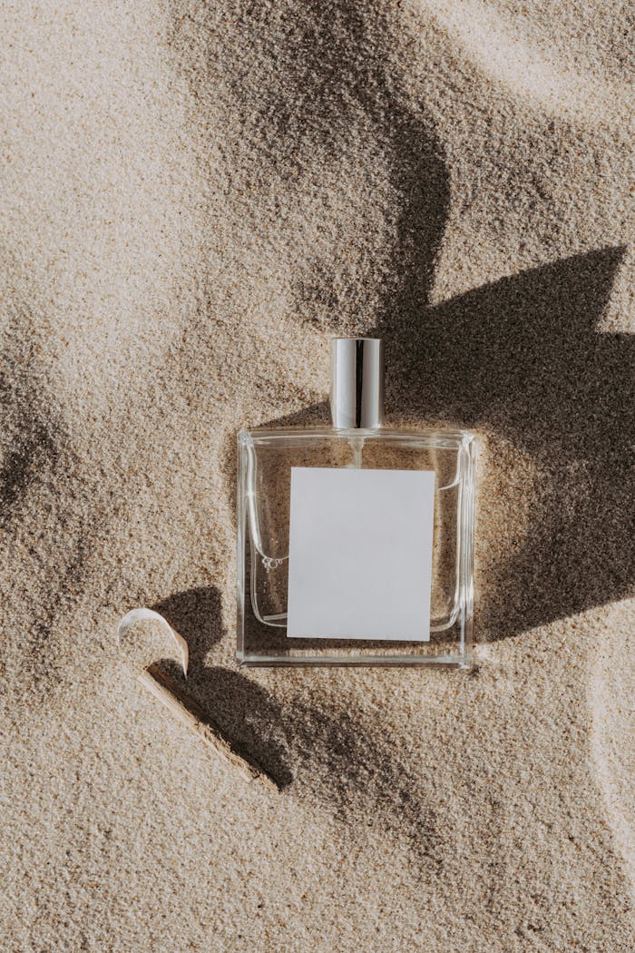 A minimalist perfume bottle with a blank label rests on sand, casting a shadow in daylight.